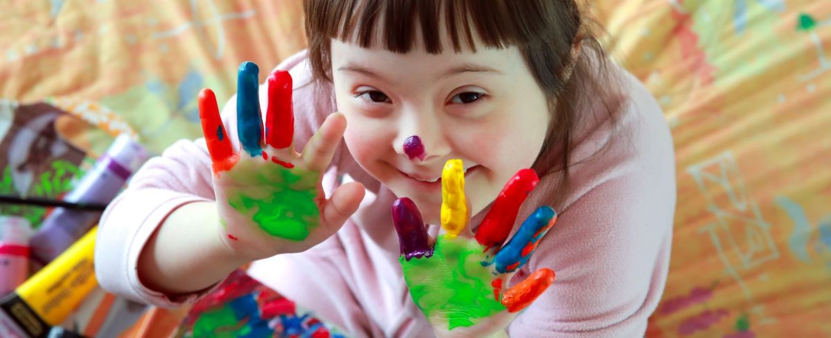 Immagine di una bambina affetta da Sindrome di Down che sorride e mostra felice le mani sporche dei colori con cui sta giocando