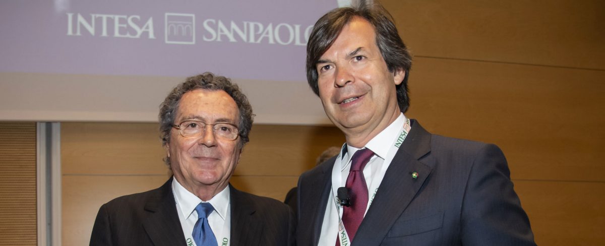 Immagine che ritrae il Presidente di Intesa Sanpaolo, Gianmaria Gros-Pietro, a fianco del CEO Carlo Messina. Vestono entrambi in abiti formali, alle loro spalle è visibile la porzione di uno schermo su cui è riprodotto il logo della Banca