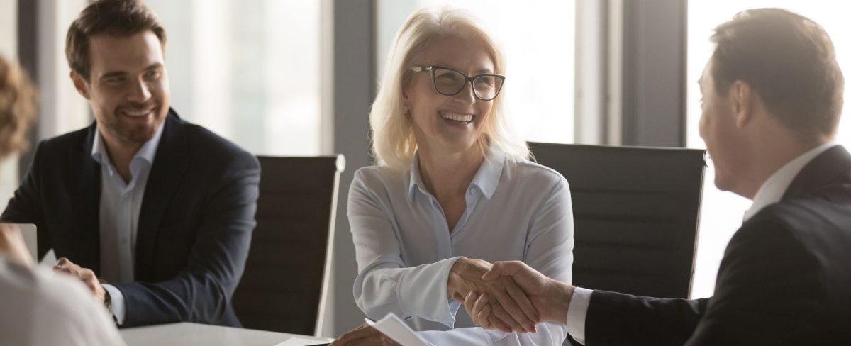 L’immagine che accompagna la News sugli strumenti a supporto dell’internazionalizzazione delle PMI italiane, grazie a un nuovo progetto che valorizza le sinergie tra la International Subsidiary Banks Division (ISBD) e la Divisione Banca dei Territori (BDT), ritrae una donna in camicia, sorridente, seduta a un tavolo riunioni e circondata da altre persone, ripresa nell'atto di stringere la mano a un uomo in giacca seduto al medesimo tavolo